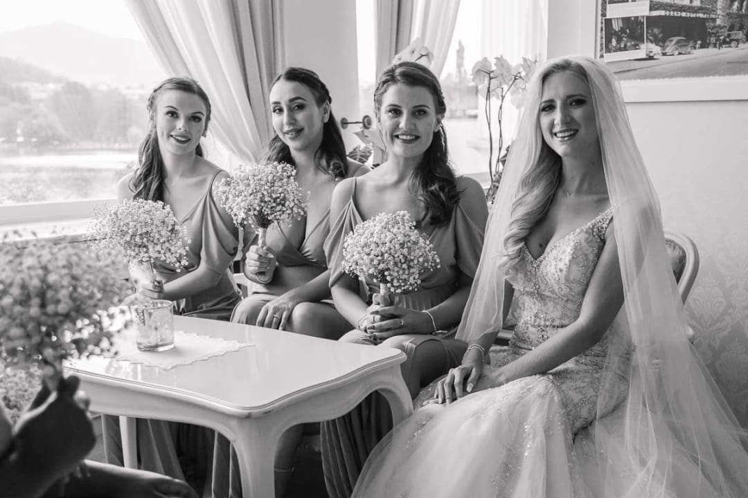 A bride sitting with her bridesmaids, each holding bouquets, inside a room with a view of the lake through the window.
