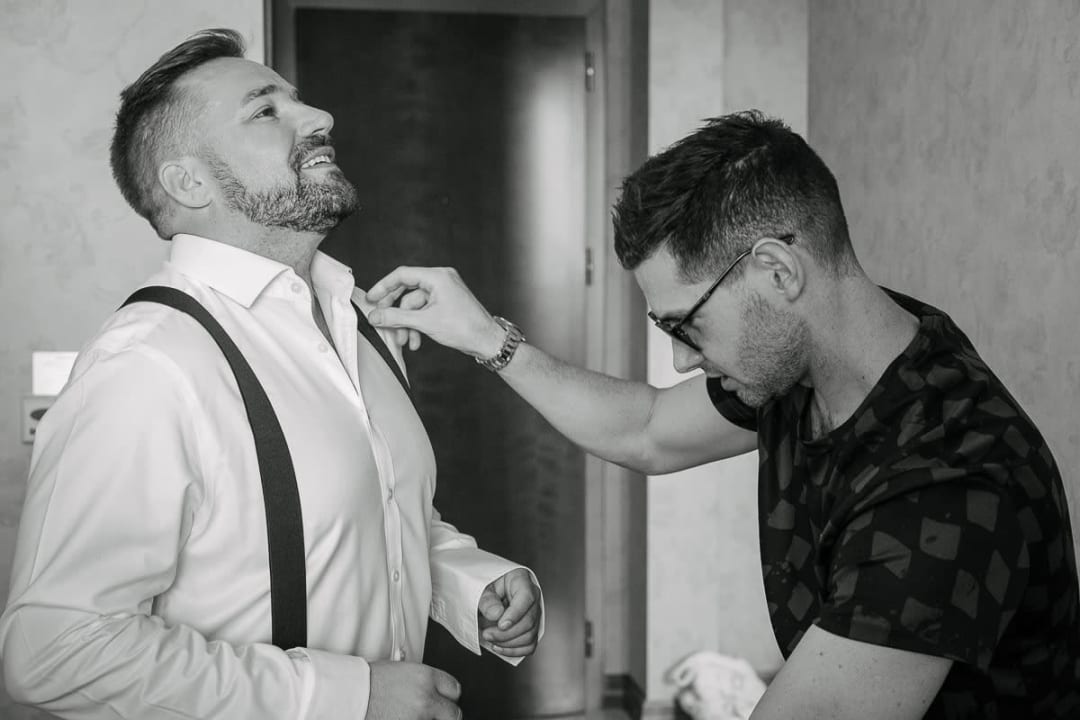 A groom is being assisted by a friend as he prepares for the wedding, adjusting his collar and suspenders.