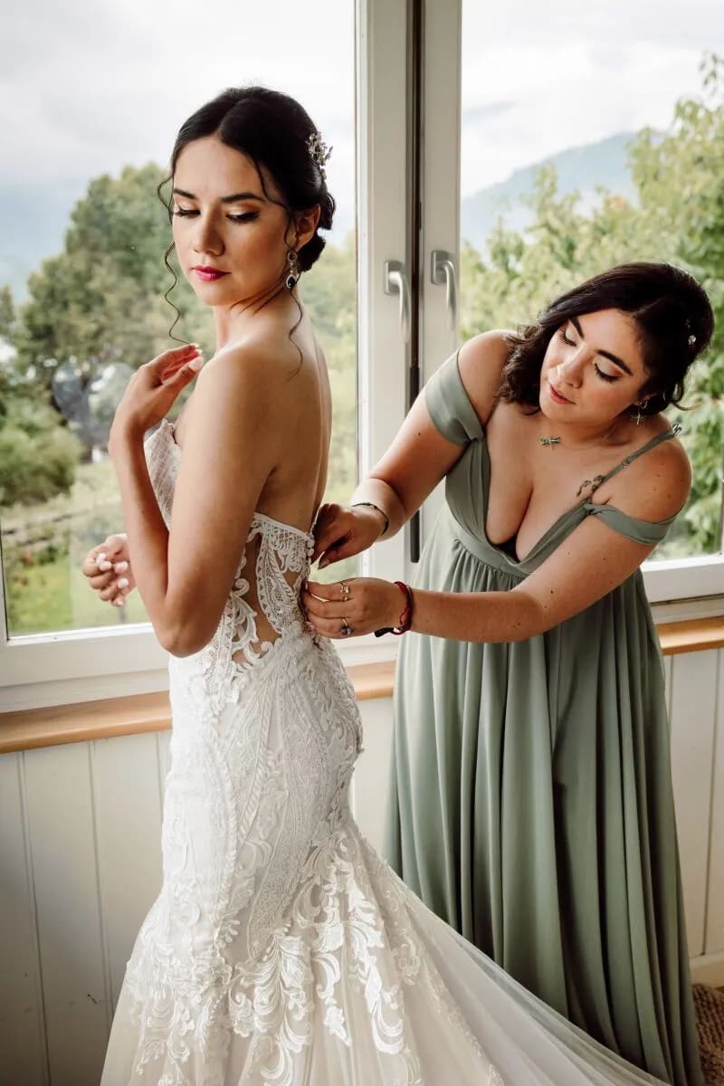 A bride standing by a window as a bridesmaid helps her with her dress. Trees and hills are visible outside.