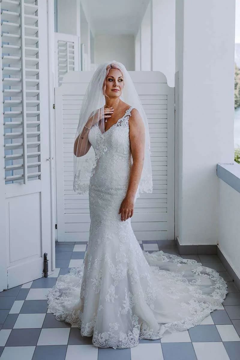 A bride in a lace wedding dress and veil stands indoors, elegantly posing in a softly lit space.