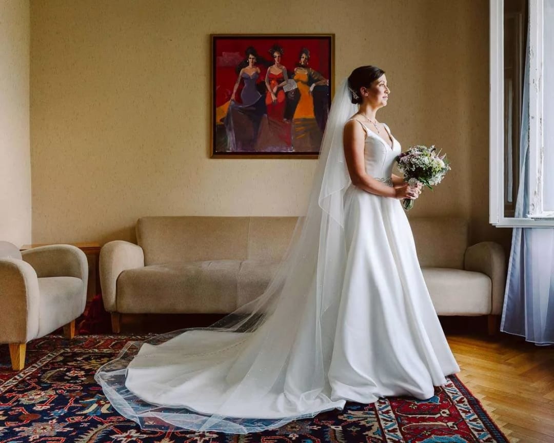 A bride in a white gown holding a bouquet, standing indoors with light streaming through a window.