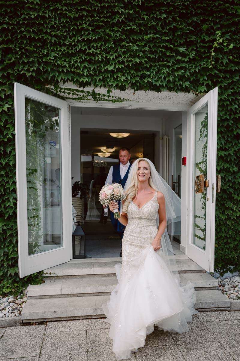 A smiling bride holding a bouquet walks through a doorway surrounded by ivy-covered walls.