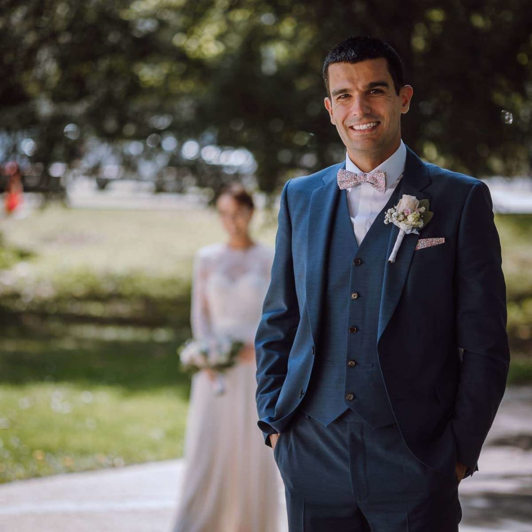 A groom in a blue suit smiling at the camera with the bride softly out of focus in the background, standing in a sunlit park.