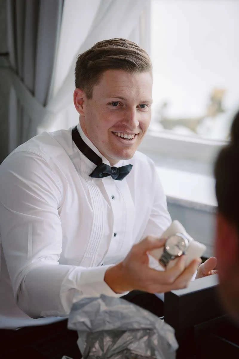 A groom wearing a white dress shirt and bow tie smiles while opening a gift, revealing a wristwatch.