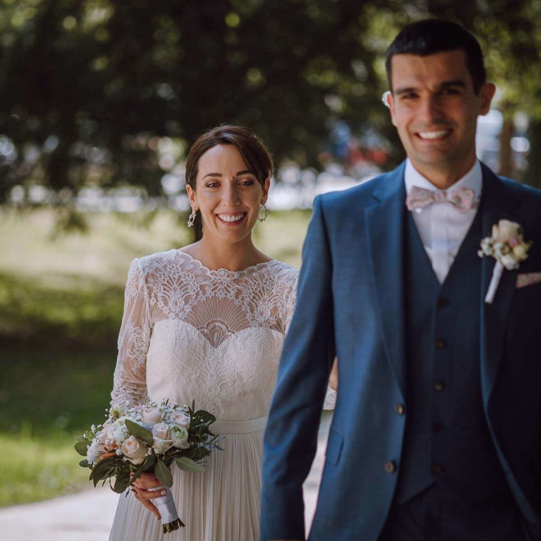 A bride holding a bouquet and smiling as she walks with the groom in a garden setting.