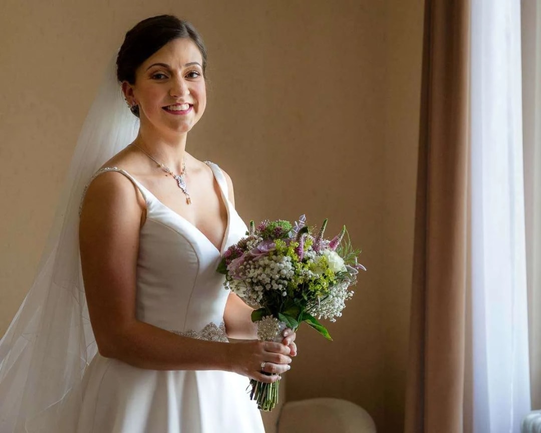 A smiling bride holding a bouquet and standing indoors, illuminated by natural light from a window.