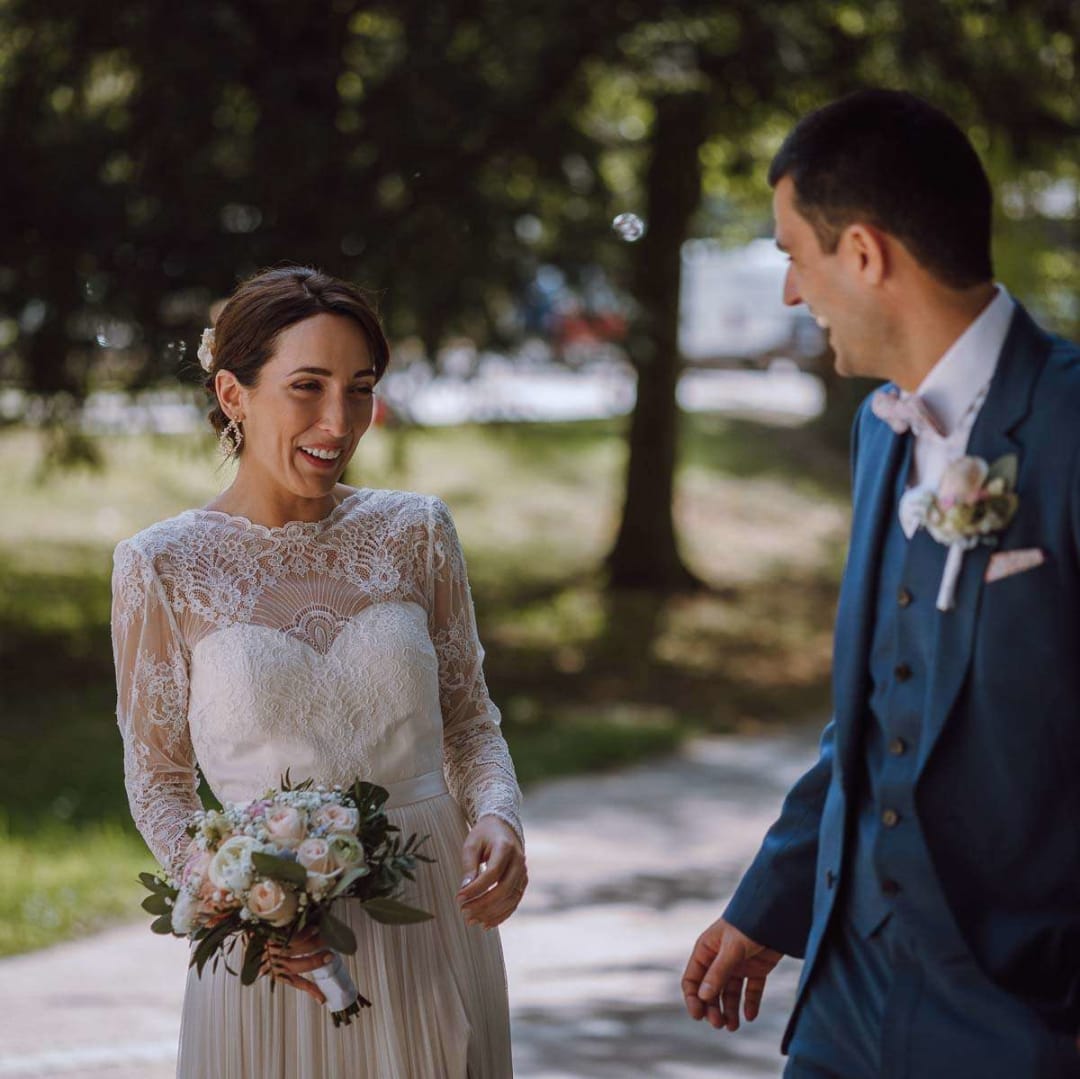A bride in a lace dress and a groom in a blue suit share a joyful moment on a shaded pathway.
