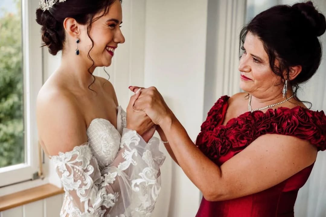 A bride in a white lace gown is smiling warmly at a woman in a red dress as they hold hands by a window.