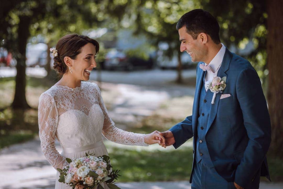 A bride and groom smiling at each other while holding hands in a garden setting.