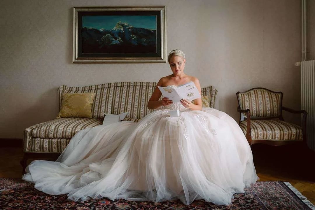 A bride in a white gown sits on a vintage sofa, reading a card in a warmly lit room.