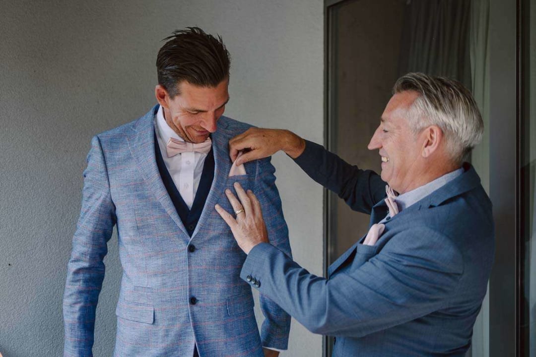 A groom smiling as an older man helps adjust his suit jacket on a wedding day.