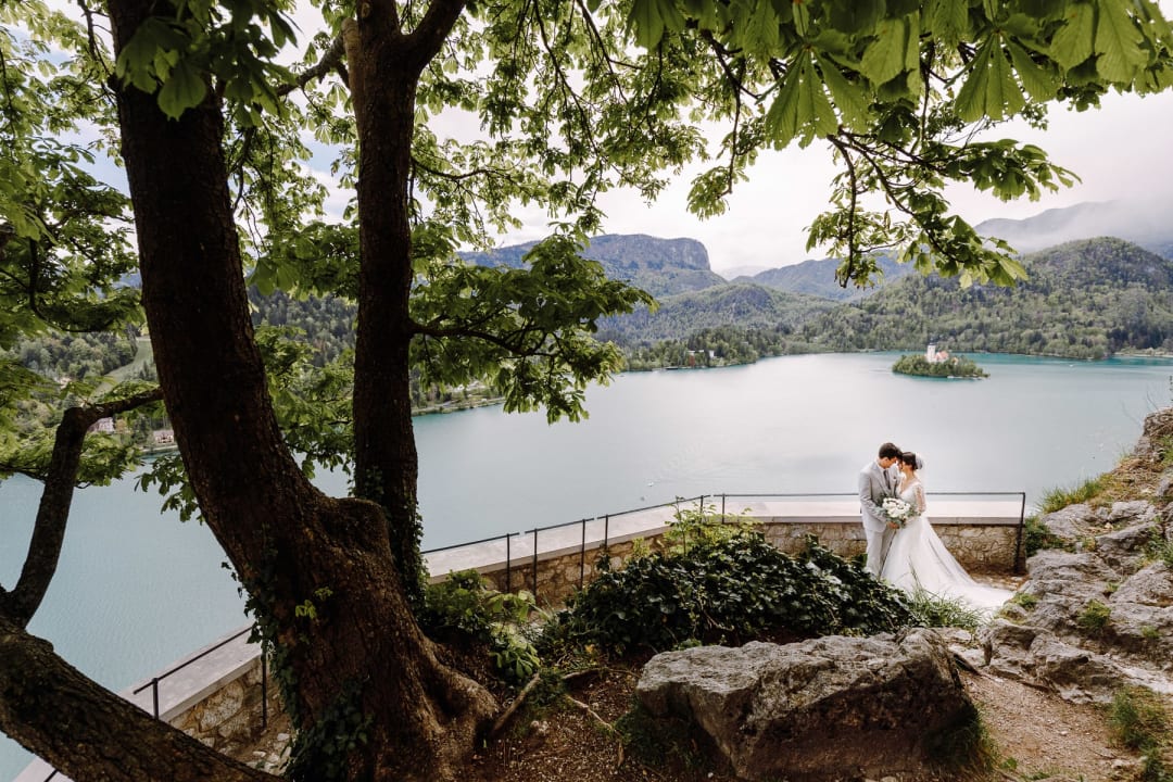 	Aerial view of Lake Bled with the tiny silhouette of a wedding couple on the castle terrace.
