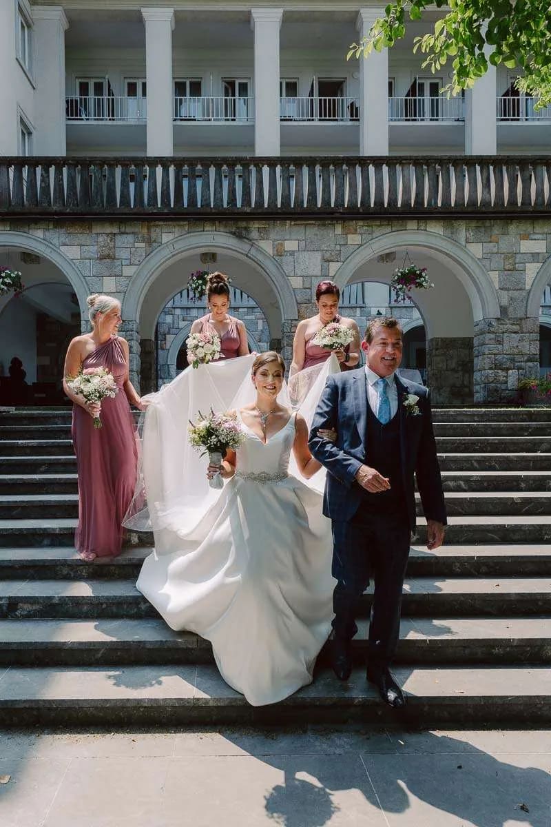 A bride accompanied by bridesmaids and a gentleman walks down the steps of a building, all smiling and holding bouquets.