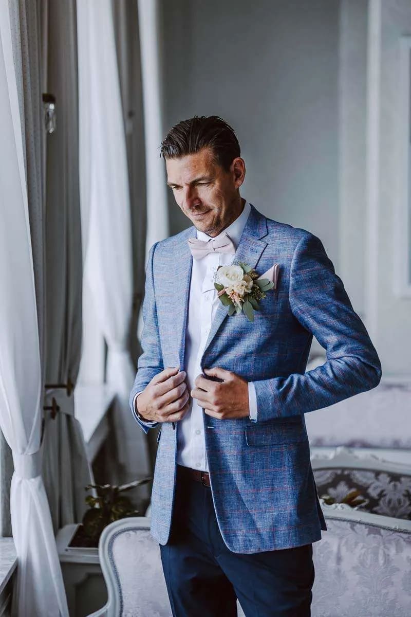 A groom adjusting his suit jacket, standing by a window, with a thoughtful expression on his face.