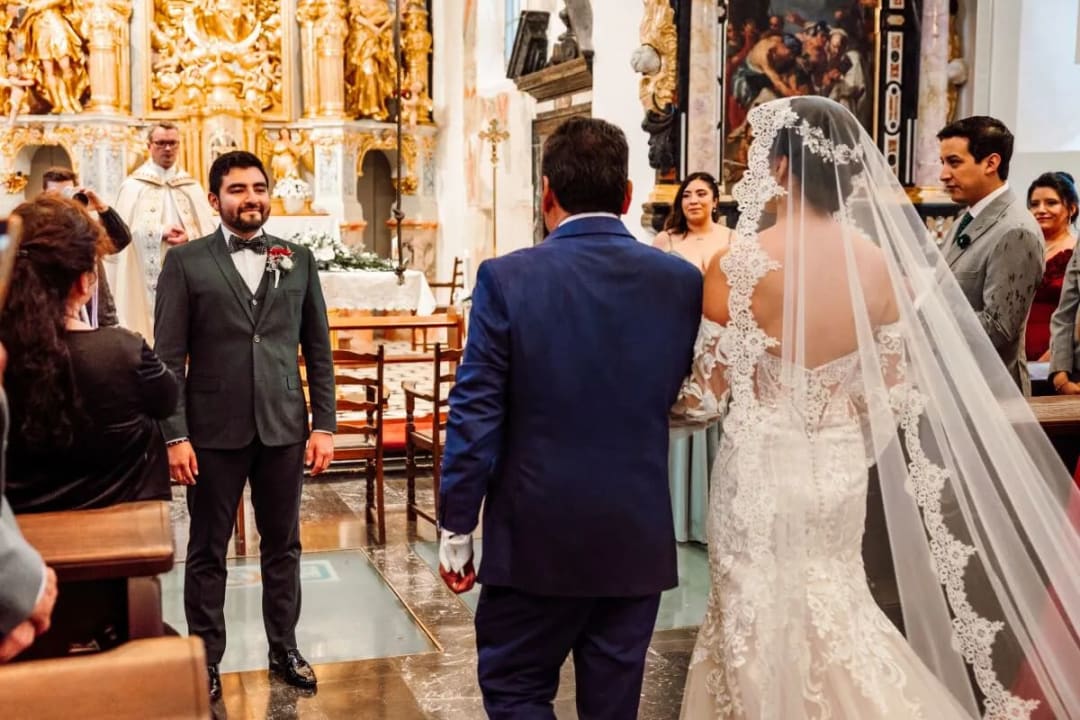 A bride walks down the aisle escorted by a man in a church, while the groom awaits at the altar.