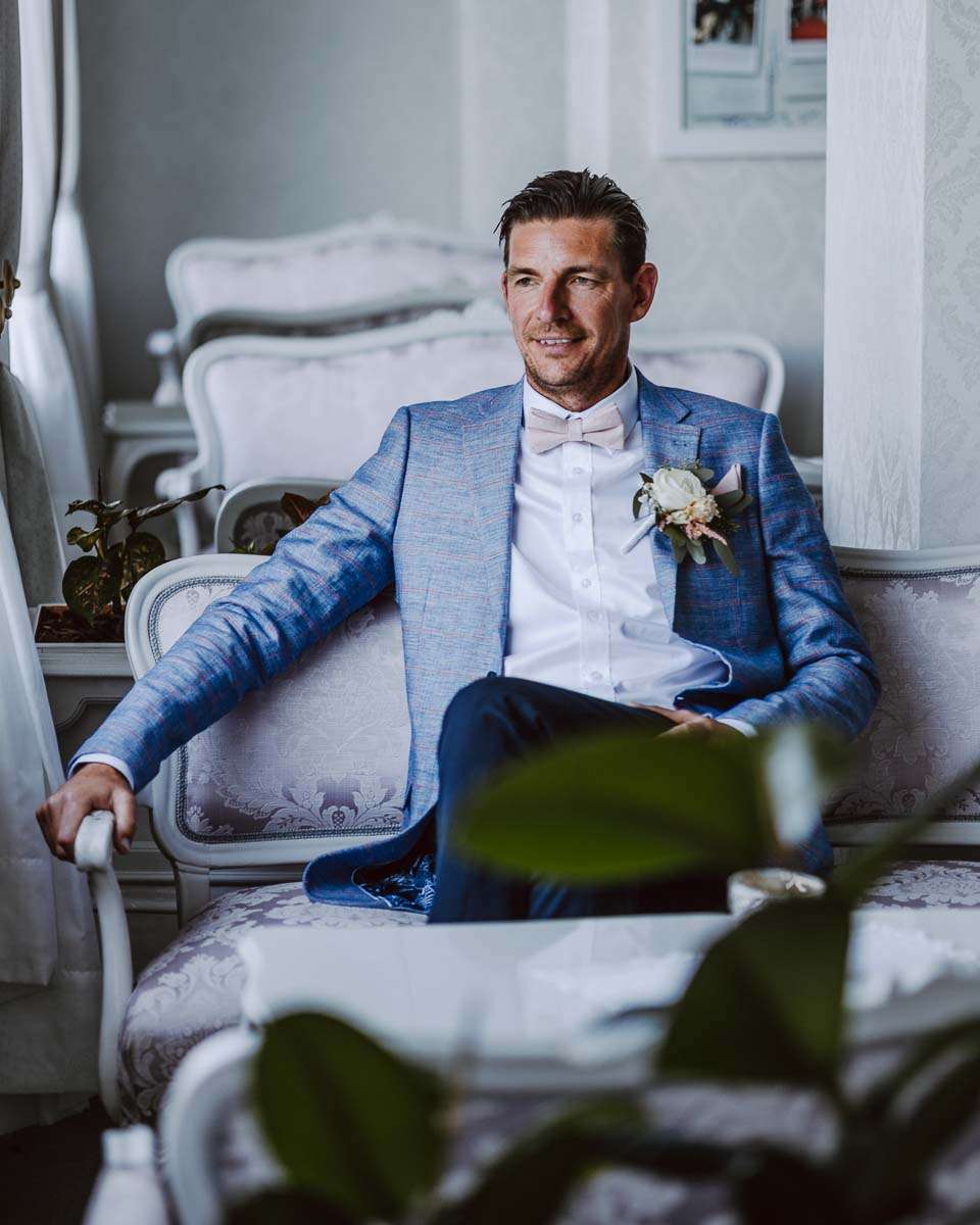 A groom in a light blue suit sitting on an elegant sofa, looking thoughtfully out the window in a well-decorated room.