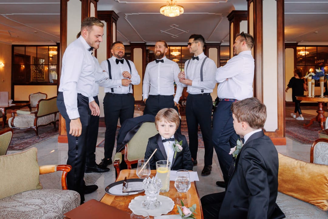 A group of groomsmen dressed in formal attire, standing and chatting in a hotel lounge, with two young boys in suits sitting at a table in the foreground.