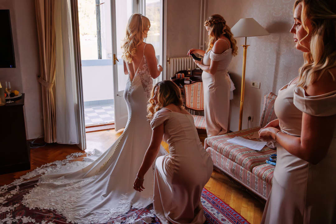 A bride in a lace wedding gown getting ready with the help of bridesmaids in a sunlit room.