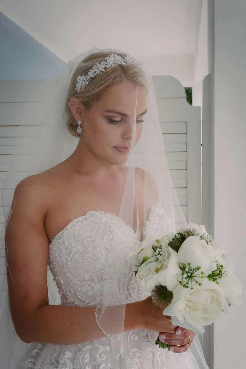 A bride holding a bouquet of white flowers, looking down, with intricate lace details on her wedding dress.