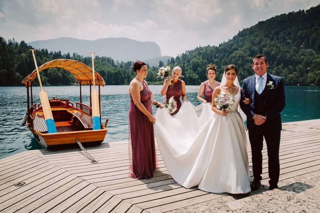 A bride and her bridal party standing on a wooden dock by Lake Bled, with a traditional wooden boat in the background.
