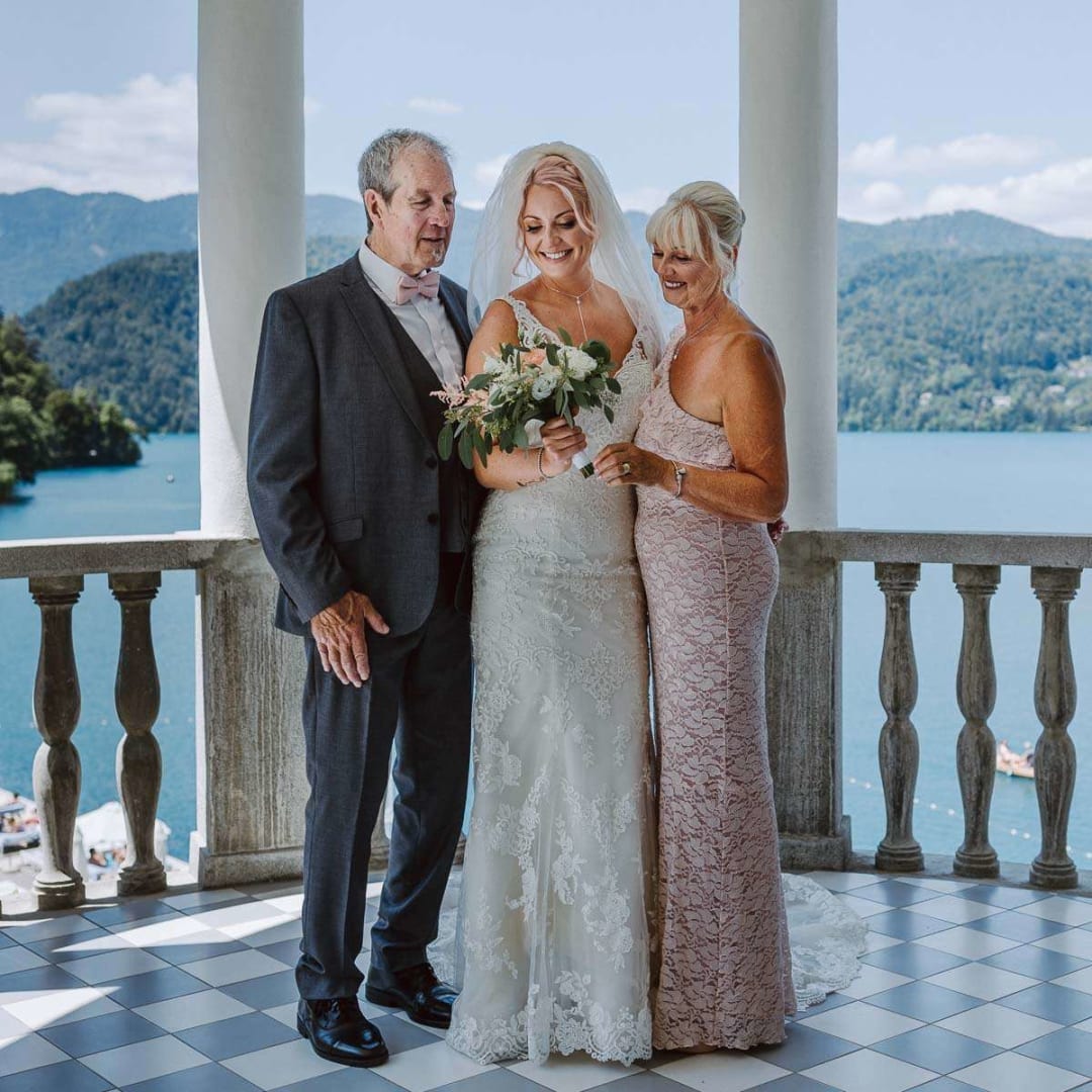 A bride in a wedding dress standing with her parents on a balcony overlooking a lake, with scenic mountains in the background.