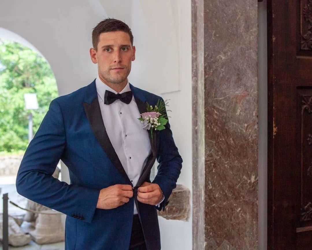 A groom in a blue suit and bow tie adjusting his jacket, standing in a stone hallway with greenery visible in the background.