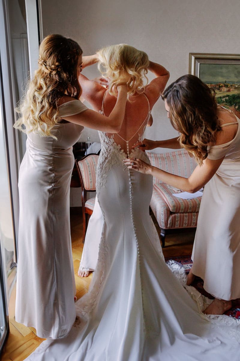 A bride is getting her wedding dress adjusted by two bridesmaids in a softly lit room.