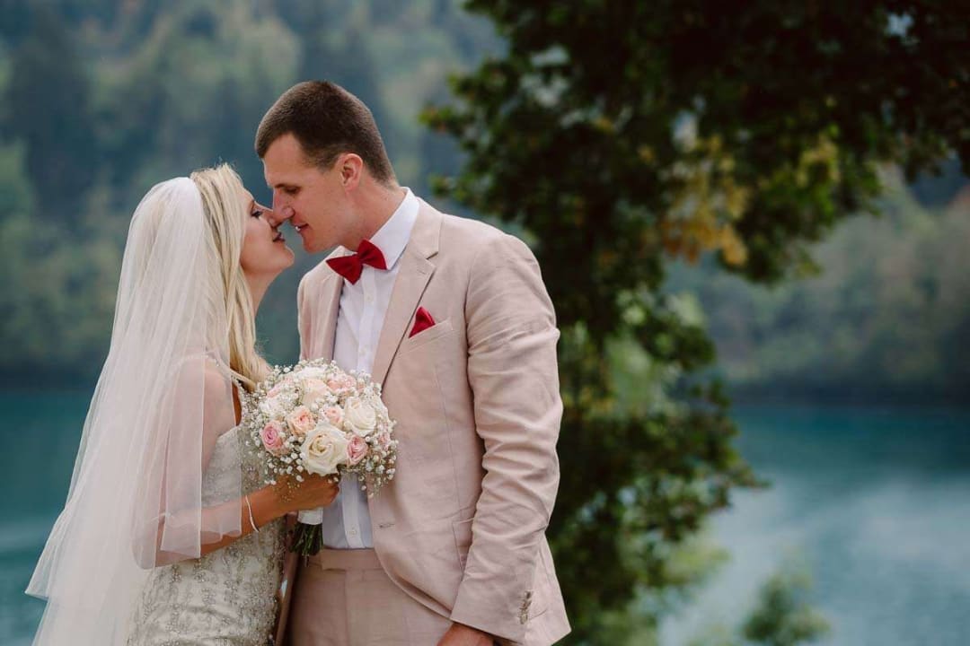 A bride and groom sharing a gentle kiss near a lake with lush greenery in the background.