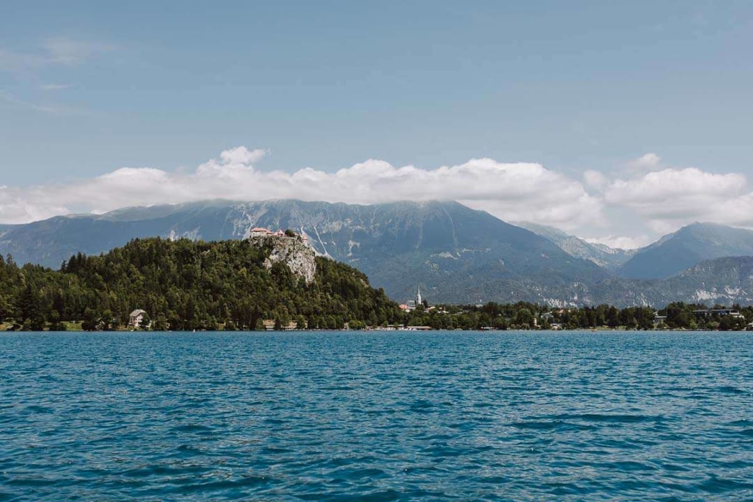 A view of Lake Bled with its clear blue water, the castle atop a hill, surrounded by lush greenery, and majestic mountains in the background under a partly cloudy sky.