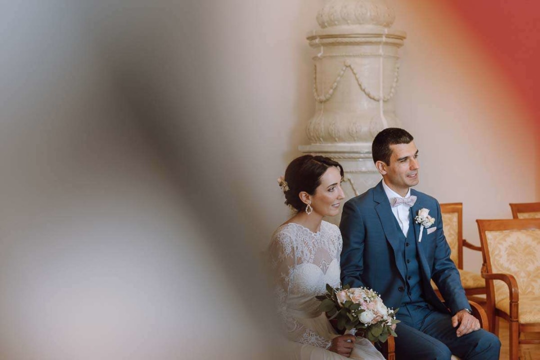A bride and groom seated inside during their wedding ceremony, with the bride holding a bouquet.