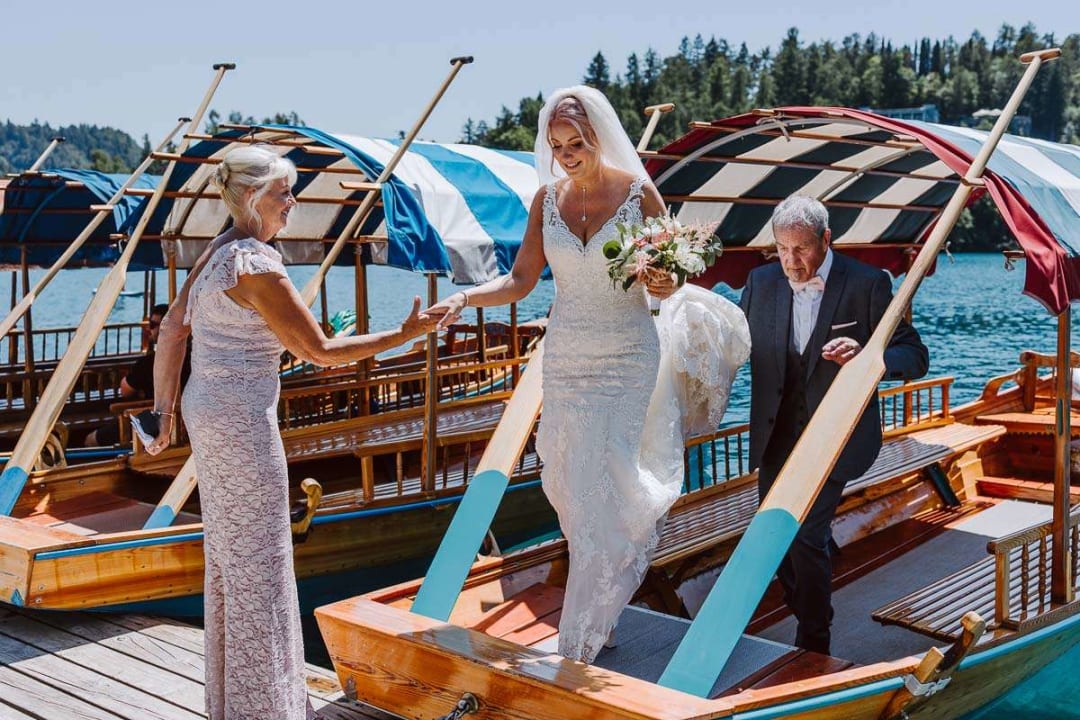 A bride stepping off a wooden boat at Lake Bled, assisted by a woman in a light dress, while an older man follows behind.