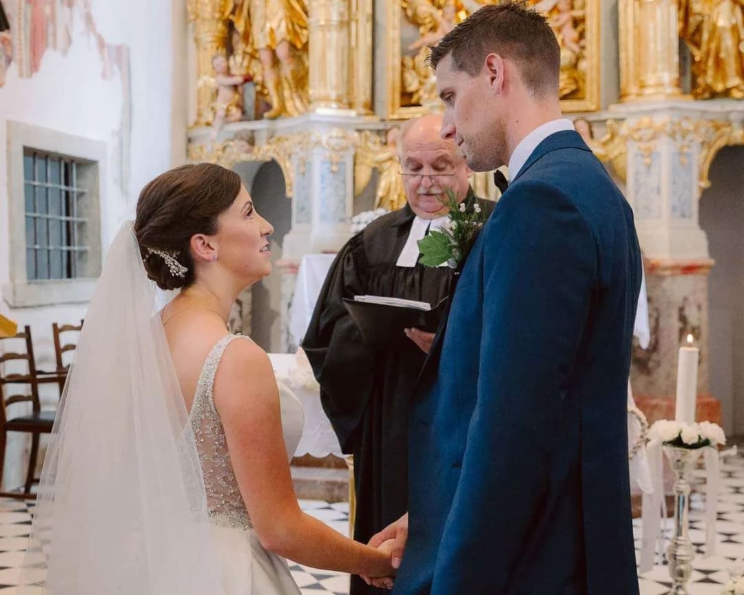 A bride and groom standing inside a beautifully decorated church, holding hands as a minister officiates the ceremony.