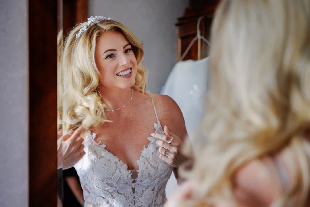 A bride smiling as she looks in the mirror while adjusting her hair during wedding preparations.