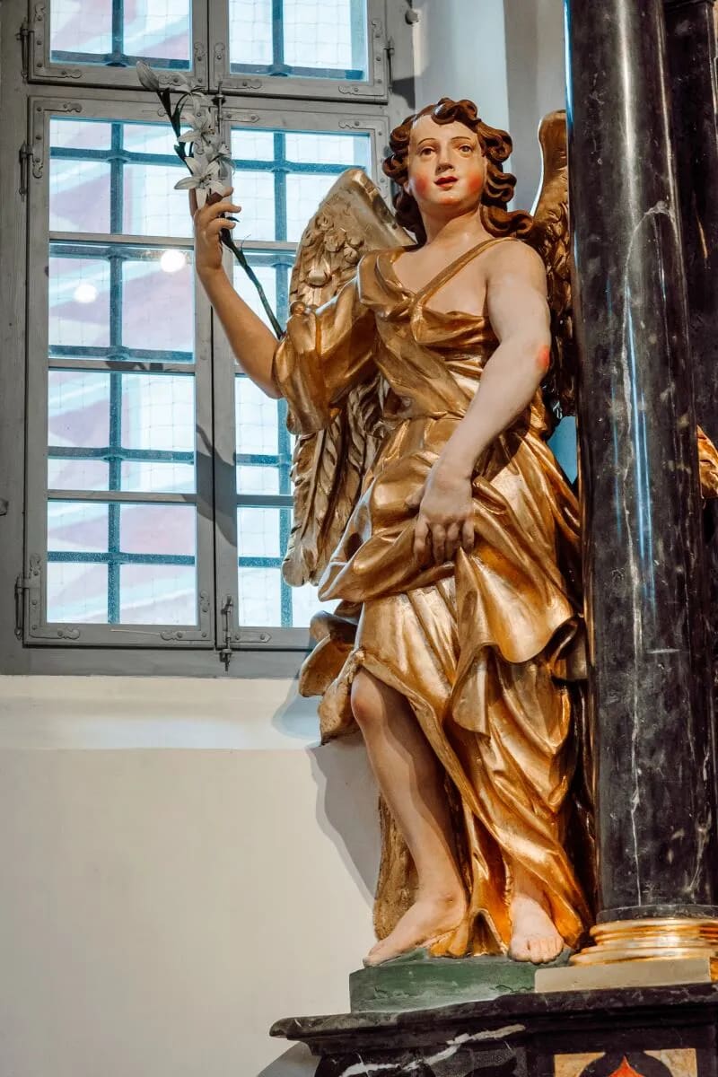 A detailed sculpture of an angel holding a flower inside a church, with a window in the background.