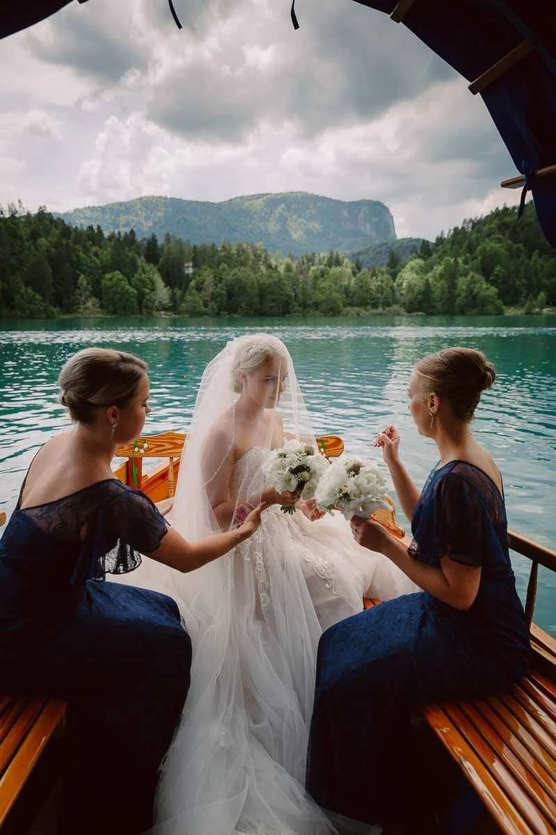 A bride sitting in a boat on a lake, surrounded by bridesmaids holding bouquets, with forested mountains in the background.