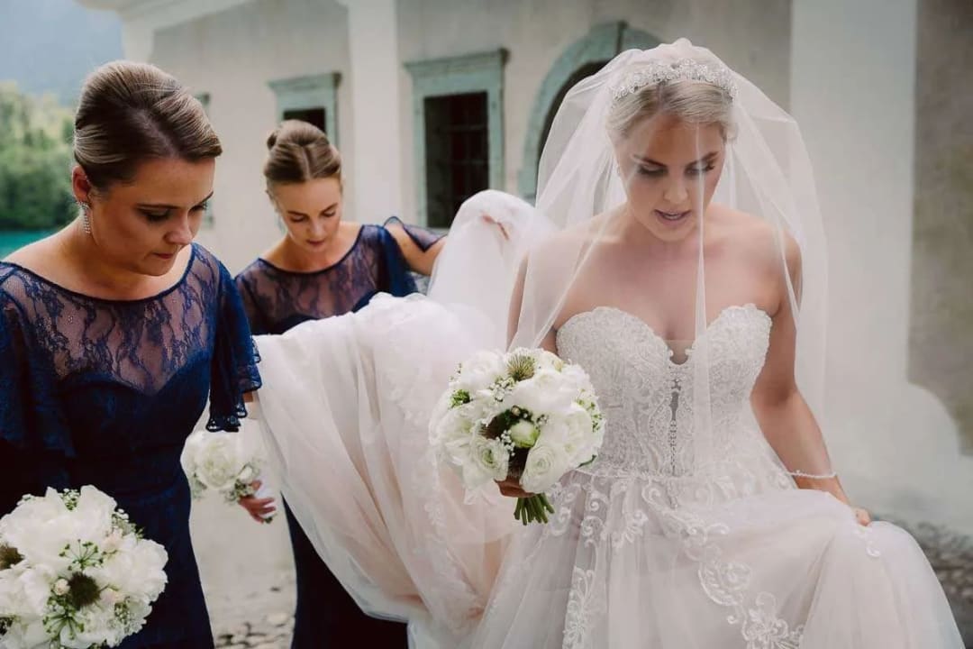 A bride in a white dress holding a bouquet of flowers is assisted by two bridesmaids in navy blue dresses, preparing for the wedding ceremony.