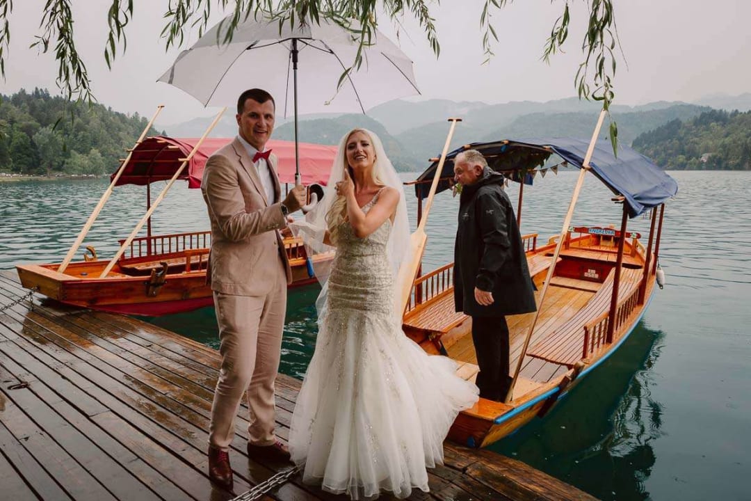 A bride and groom pose under an umbrella on a wooden dock with traditional boats and a scenic lake in the background.