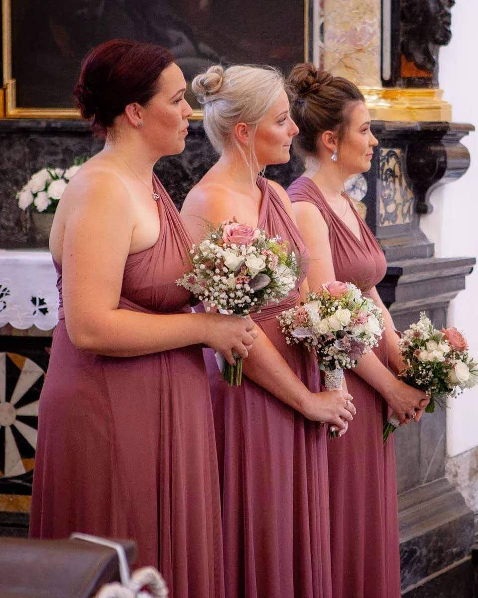 Three bridesmaids in matching dresses holding bouquets of flowers during a wedding ceremony in a decorated venue.