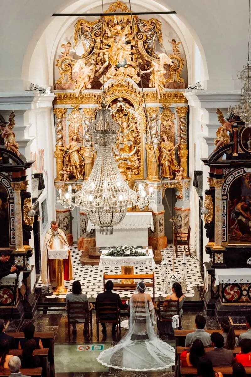 A bride and groom sitting in a beautifully ornate church during their wedding ceremony, featuring intricate gold decor and a large chandelier.