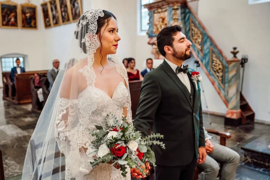 A bride and groom standing together in a church during their wedding ceremony, with the bride holding a bouquet of red and white flowers.