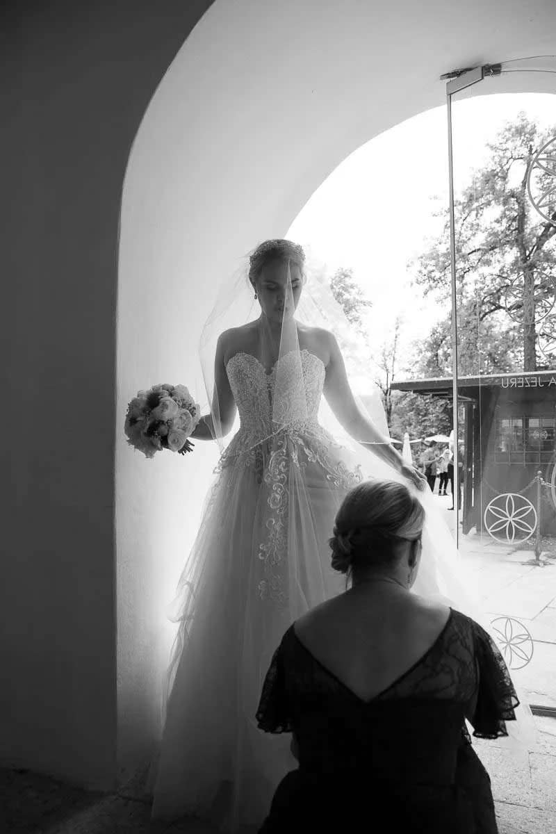 A bride in an elegant gown holding a bouquet, standing under an archway with a woman adjusting her dress.