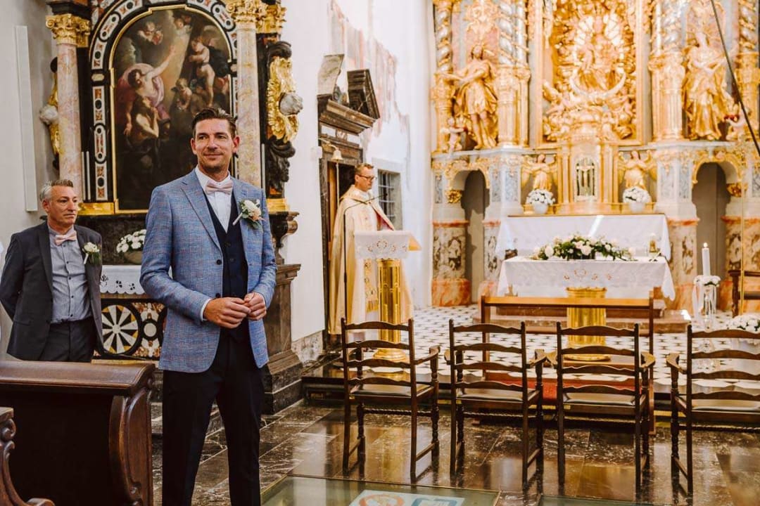 A groom stands at the front of an ornately decorated church with a priest in the background, ready for the wedding ceremony to begin.