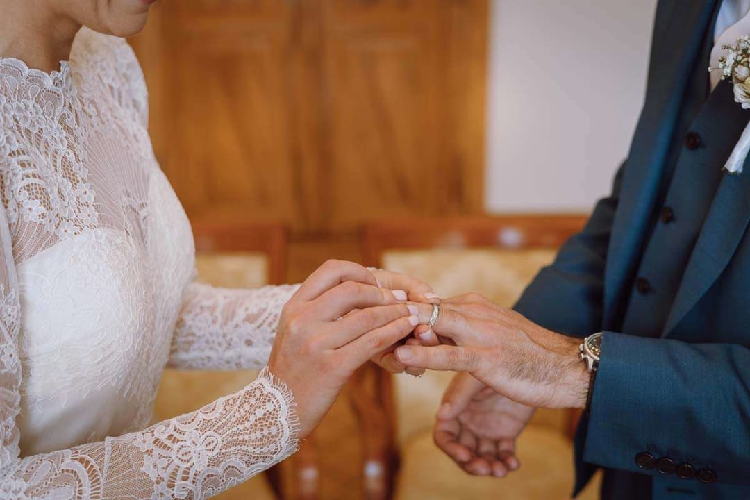 A bride placing a wedding ring on the groom's finger during a ceremony indoors.