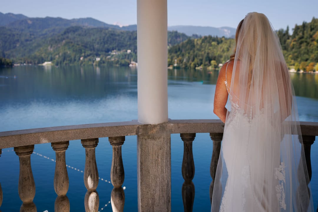 A bride stands on a balcony, overlooking the serene waters of Lake Bled with forested mountains in the backdrop.