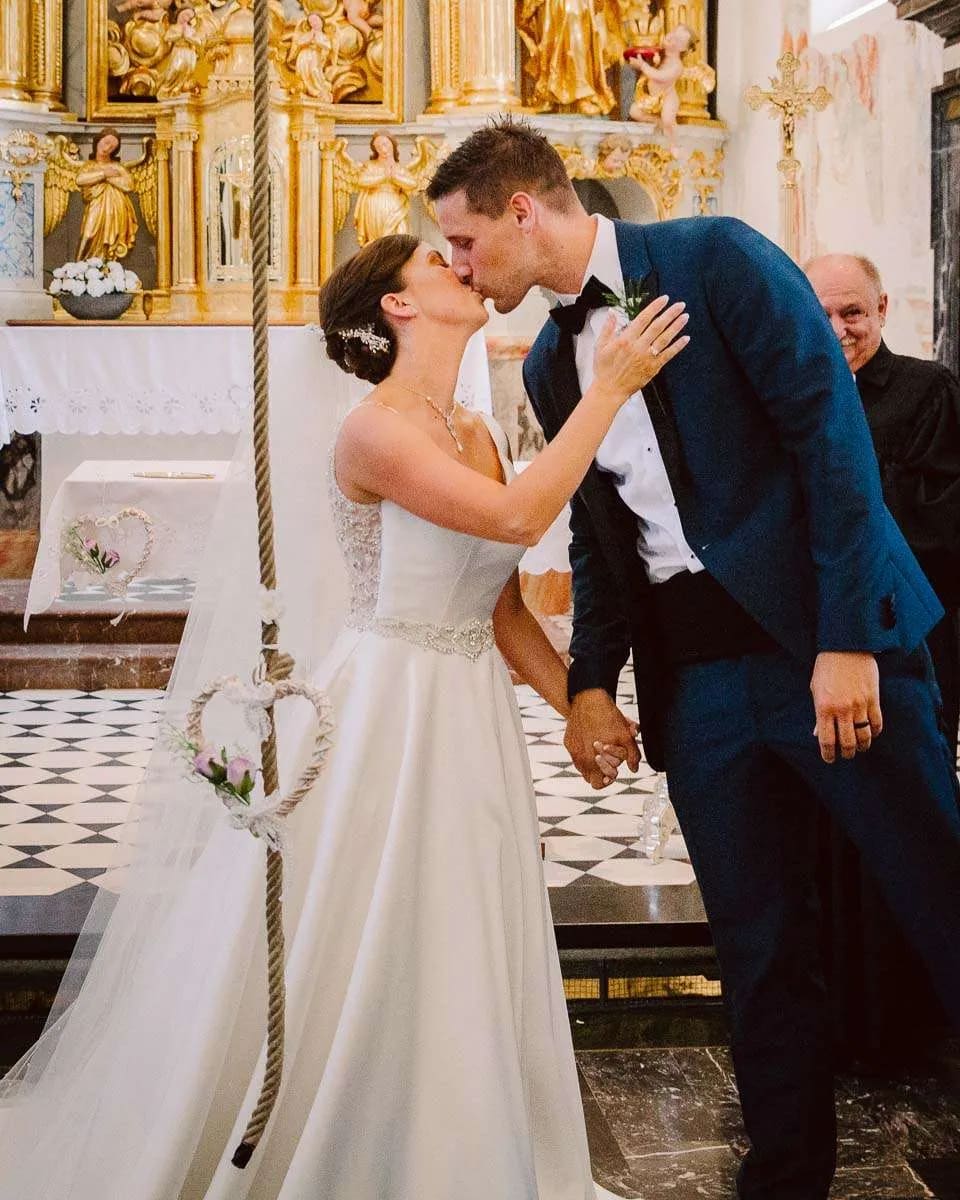 A bride and groom share a kiss inside a beautifully decorated church, holding hands in front of the altar.