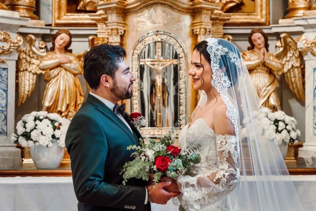 A bride and groom standing together inside a church, sharing a loving gaze with floral arrangements and golden angel statues in the background.