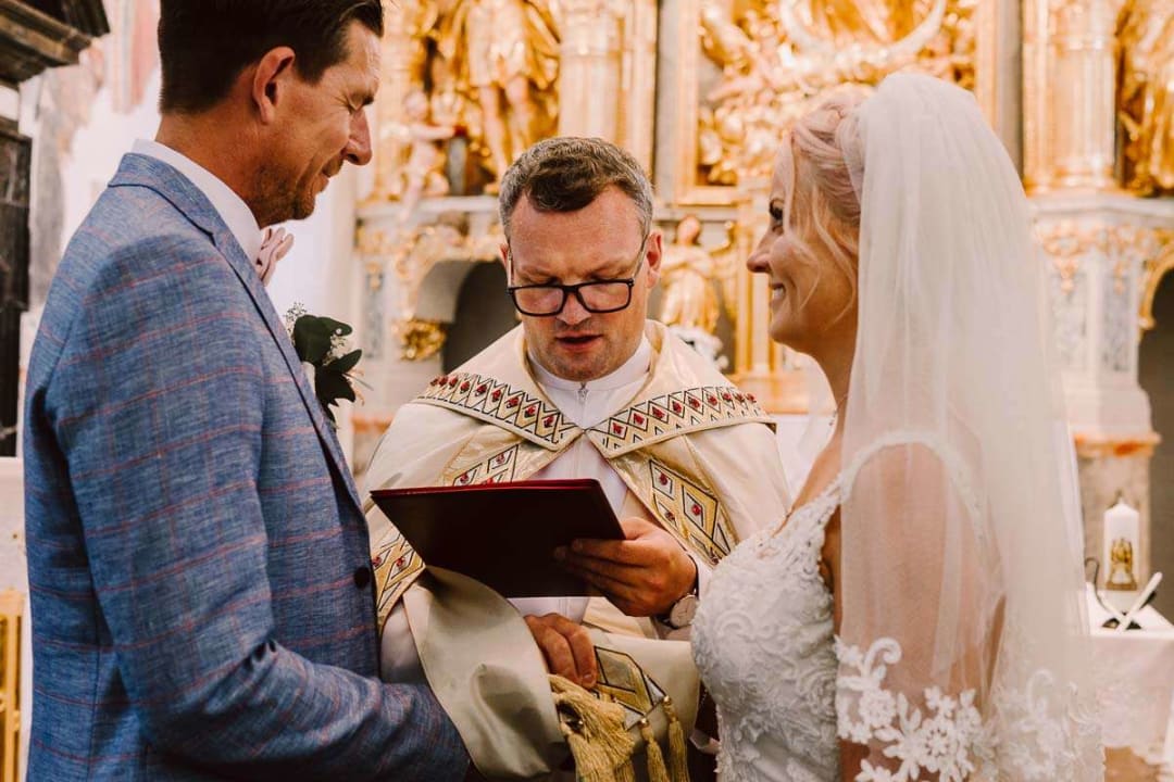 A bride and groom exchanging vows during a wedding ceremony, with a priest reading from a book in an ornate church setting.