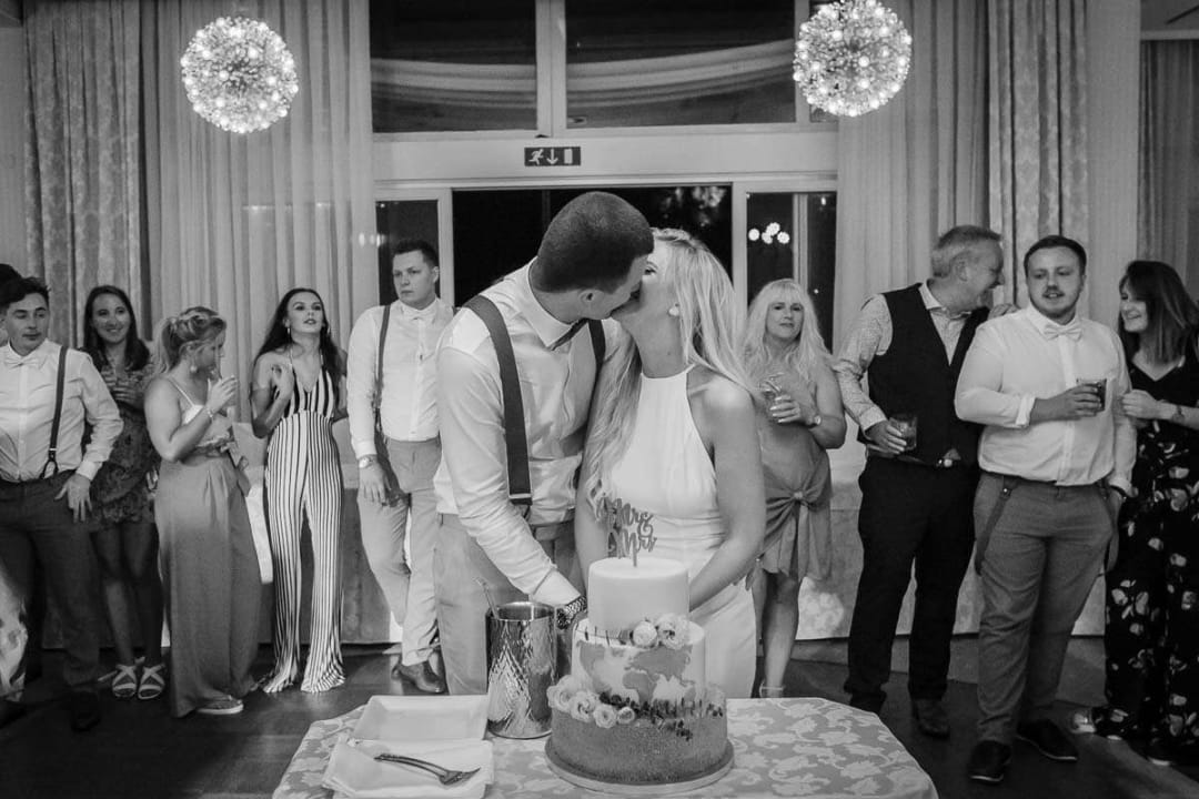 A bride and groom share a kiss during their cake cutting ceremony, surrounded by guests at the wedding reception.