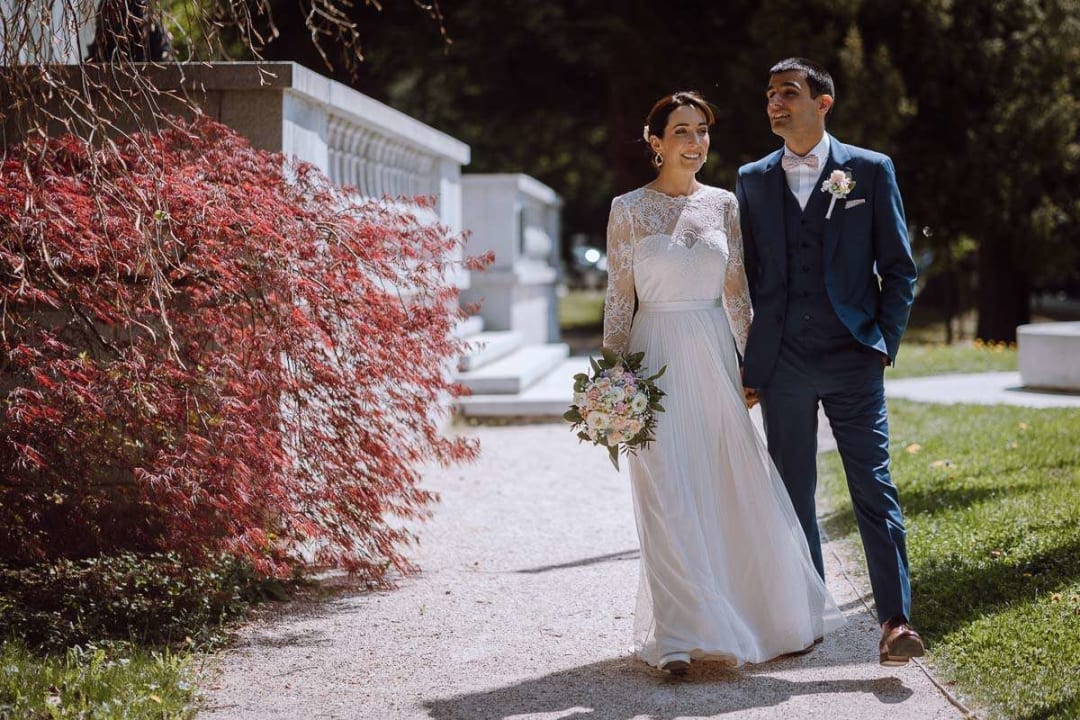 A bride and groom walking together down a garden path, surrounded by lush greenery and vibrant red foliage.