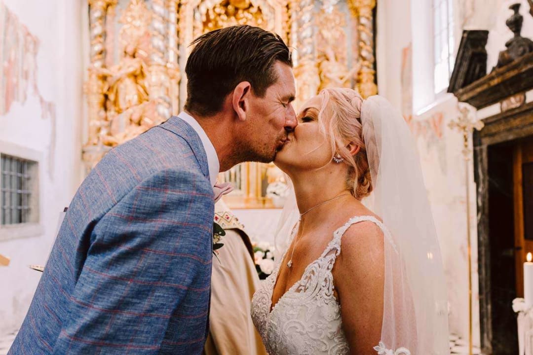 A bride and groom sharing a kiss inside a beautifully decorated church with ornate golden accents in the background.